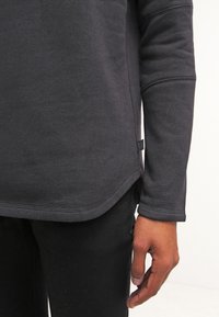 Camiseta de manga larga gris oscuro con un dobladillo redondeado, tela texturizada, detalles de costura visibles y una pequeña etiqueta en el lateral.