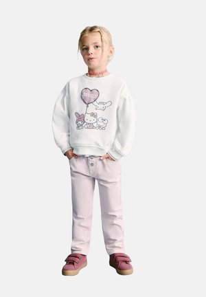 Jeune fille aux tresses blondes portant un sweat-shirt blanc Hello Kitty, un pantalon rose clair, des baskets roses et un collier rose, debout avec les mains dans les poches.