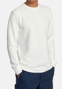 Chemise blanche à manches longues avec un col rond, dotée d'un tissu texturé. Portée avec un pantalon bleu marine, les mains dans les poches.