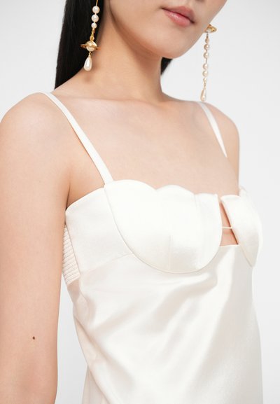 Žena v bielych saténových šatách s tenkými ramienkami a výstrihom v tvare srdca, doplnená zlatými a perlovými náušnicami na visiace slzy.