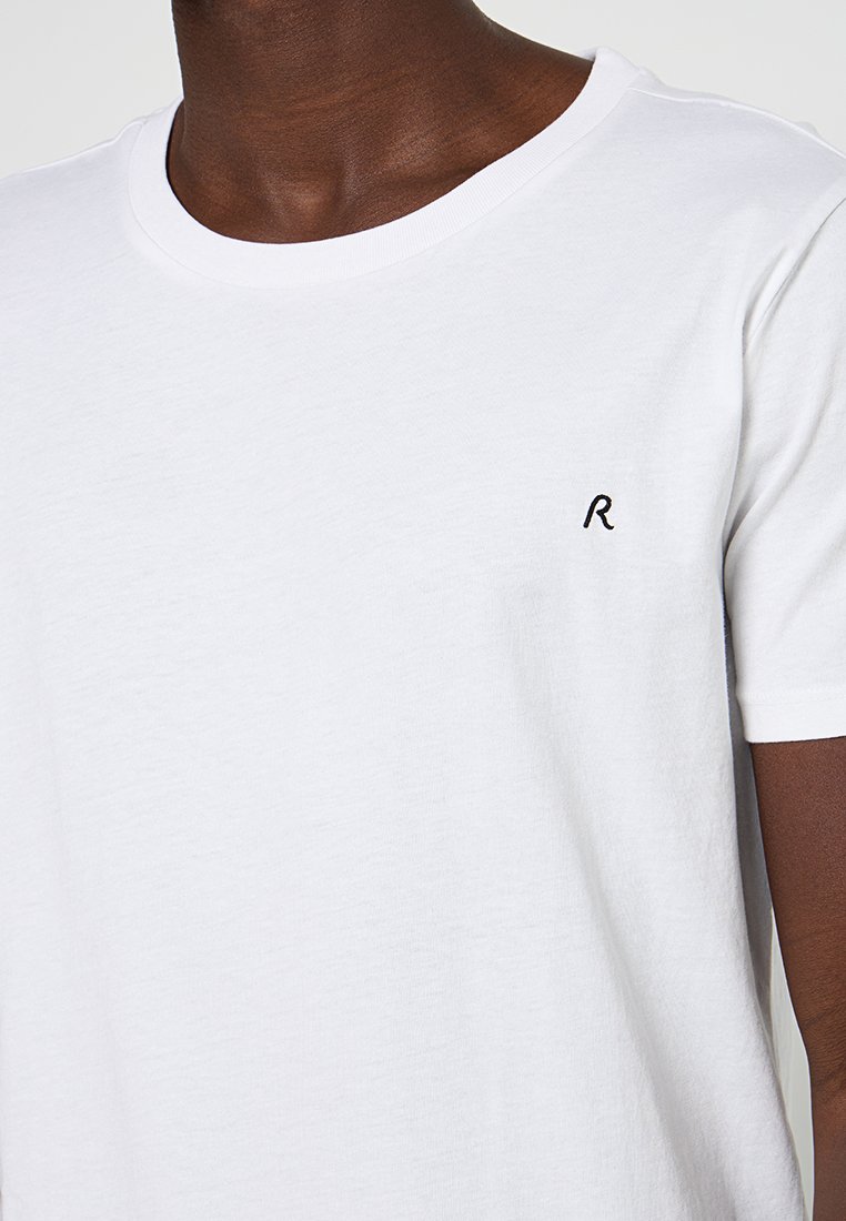 Biała bawełniana koszulka z okrągłym dekoltem, krótkimi rękawami i małym czarnym haftowanym "R" po lewej stronie klatki piersiowej. Gładka faktura materiału.