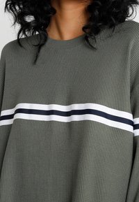 Ribbad grön tröja med rund hals, med horisontella vita och marinblå ränder över bröstet. Mjuk textur, avslappnad passform.