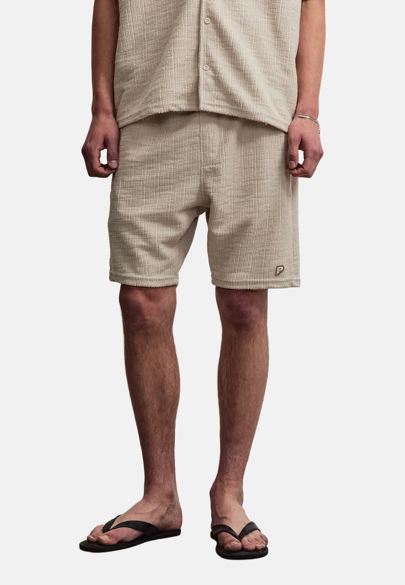 Homme portant un short beige texturé et une chemise assortie à manches courtes, tongs noires, debout devant un fond uni.