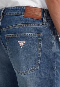 Plave traperice s kožnom etiketom Guess Jeans i trokutastim crvenim i bijelim logotipom na stražnjem džepu. Ima vidljive šavove.