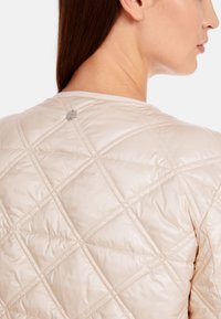 Hellbeige gesteppte Jacke mit einem Rautenmuster, glatter Textur und einem runden Knopfdetail am hinteren Halsausschnitt.