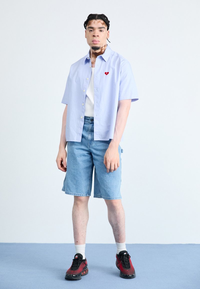 Chemise à manches courtes de couleur bleu clair avec un logo en forme de cœur rouge, portée sur un débardeur blanc ; associée à des shorts en denim bleu et des baskets noires.