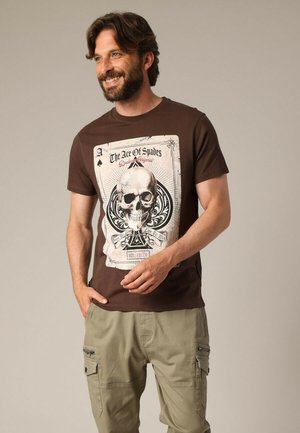 Bruine t-shirt met een grafische print van een schedel en speelkaarten ontwerp. Korte mouwen en gemaakt van zachte katoen.