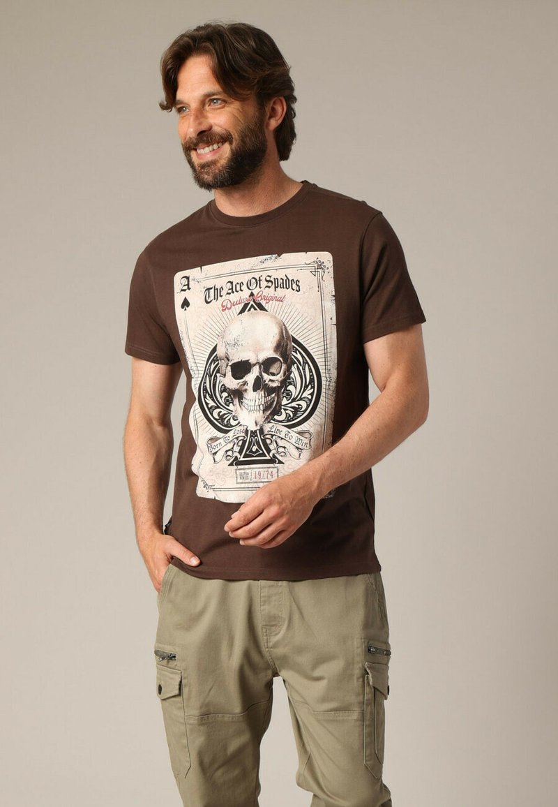 T-shirt marron avec un imprimé graphique représentant un crâne et un motif de carte à jouer. Manches courtes et matière en coton douce.