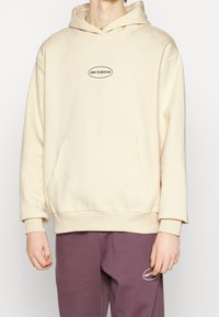 Felpa crema con tasca frontale e logo ricamato. Presenta un cappuccio con coulisse e polsini a costine, abbinata a pantaloni completi viola.