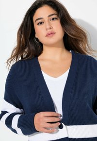 Tmavě modrý pletený kardigan s bílými pruhy, designem s výstřihem do V, zapínáním na knoflíky a texturovanou látkou. Nosený přes bílé tričko.