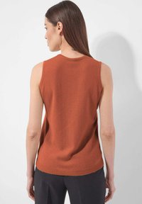 Eine Frau mit langen braunen Haaren trägt ein ärmelloses, rostfarbenes Oberteil und dunkle Hosen. Sie steht vor einem schlichten weißen Hintergrund und dreht sich weg.