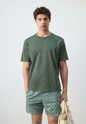 Jonge man draagt een groen t-shirt en een korte broek met patroon, houdt een beige tote tas vast en staat tegen een effen witte achtergrond.