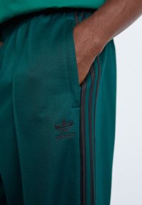 Adidas mørkegrønne sweatpants med sorte sidelister og logo, lavet af tekstureret stof, med sidelommer og elastisk talje.
