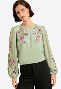 Gaiši zaļa blūze ar ziedu izšuvumiem rozā un purpura krāsā. Ar puffētiem caurspīdīgiem piedurknēm un apaļu kaklu. Saskaņota ar melnām biksēm.