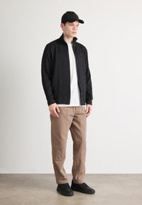 Giacca nera con zip e colletto alto, abbinata a una t-shirt bianca e pantaloni marroni. Sneakers nere completano il look.