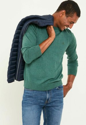 Grüner Strickpullover mit gerippten Bündchen und Saum, getragen mit blauen Jeans. Eine marineblaue Daunenjacke ist über eine Schulter gelegt.