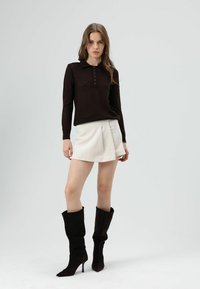 Bruine trui met textuur, kraag en knoopsluiting, gecombineerd met een witte minirok en zwarte kniehoge laarzen met hak.