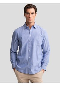 Bărbat tânăr cu păr castaniu scurt, purtând o cămașă deschisă la culoare cu nasturi și pantaloni bej, stând pe un fundal simplu gri.