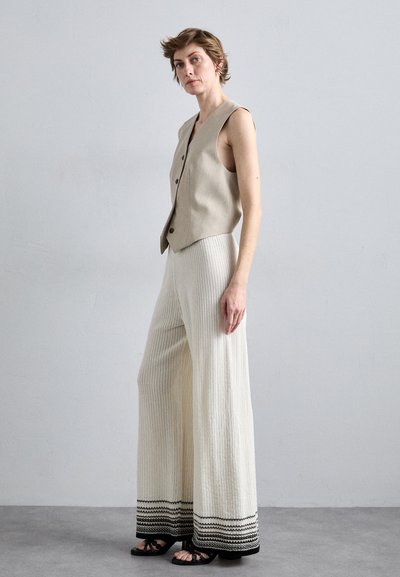 Beige mouwloze vest met knopen aan de voorkant, gecombineerd met wijde witte textuurbroeken met zwarte gestreepte accenten aan de zoom. Zwarte sandalen.