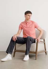 Polo rose avec un col contrastant, associé à un jean gris foncé et des baskets blanches. Modèle assis sur une chaise en bois de couleur claire.