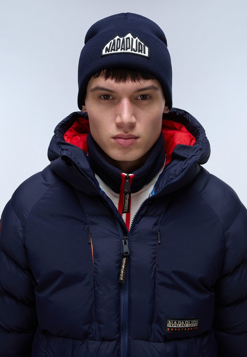Giubbotto imbottito navy con colletto alto, tasche con zip e interno rosso. Abbinate a un berretto in maglia navy con patch logo bianco.