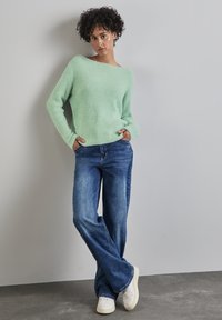 Suéter de punto texturizado en verde menta con escote amplio, combinado con jeans de campana azules y zapatillas blancas. La modelo está de pie frente a una pared neutra.