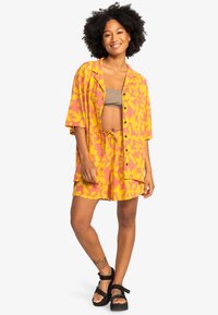 Camisa de manga corta con botones y pantalones cortos a juego en rosa con un patrón floral amarillo. La tela tiene una textura ligera y suave. Sandalias negras.