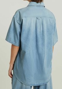 Ljusblå denimskjorta med korta ärmar, bakveck, rundad nederkant och subtil textur. Har en klassisk krage och en avslappnad design.
