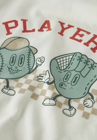 Camiseta blanca con diseños de dibujos animados de una pelota de béisbol y un guante, con el texto rojo "JUGAR" arriba y un patrón de cuadros marrones abajo.