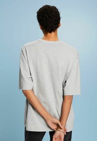 Grå t-shirt med kort ärm gjord av mjukt tyg med lös passform och rund halsringning, sedd bakifrån mot en ljusblå bakgrund.