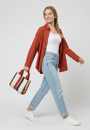 Jauna moteris vaikšto, dėvi rūdžių spalvos atvirą marškinių viršutinę dalį, baltą palaidinę, šviesiai mėlynas džinsus, baltus sportbačius, dešinėje rankoje neša dryžuotą rankinę.