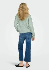 Pull tricoté vert clair avec motifs de cœurs, associé à un jean bleu raccourci et des chaussures noires sans lacets. Cheveux coiffés en ondulations lâches.