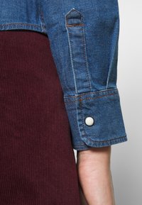 Denimskjortärm med blått tyg, orange sömnad och en vit tryckknapp. Bakgrunden har en vinröd ribbad struktur.