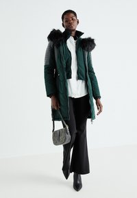 Zaļš polsterēts parka ar mākslīgā kažoka apkakli, ar siltuma šuvēm un pelēkiem akcentiem. Saskaņots ar baltu krekli, melnām biksēm un pelēku pleca somu.