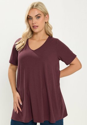 Frau mit blonden Haaren, die ein locker sitzendes, kurzärmeliges, geripptes, dunkelviolettes V-Ausschnitt-Oberteil trägt und posiert, wobei eine Hand auf der Hüfte ruht und die andere in der Tasche steckt.