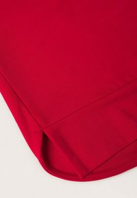 Gros plan d’un ourlet de manche en tissu rouge avec une petite fente latérale et des coutures soignées sur un fond clair.