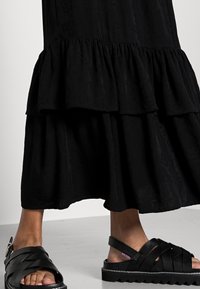 Robe maxi noire à volants en tissu texturé, avec des ruffles superposées, associée à des sandales noires à lanières avec une semelle épaisse et des détails de boucle.