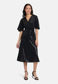Robe noire à sequins avec un design portefeuille, manches courtes et taille cintrée, tombant en dessous du genou. Assortie à des talons noirs à lanières.