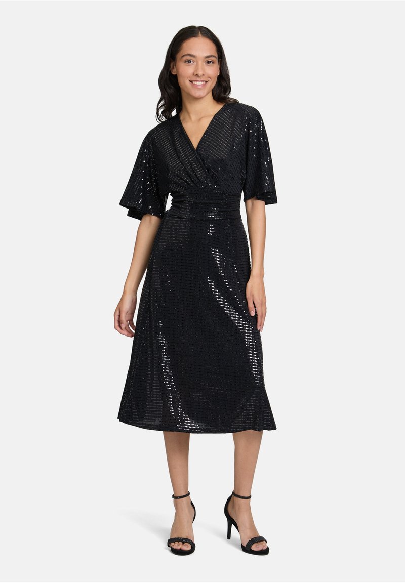 Robe noire à sequins avec un design portefeuille, manches courtes et taille cintrée, tombant en dessous du genou. Assortie à des talons noirs à lanières.