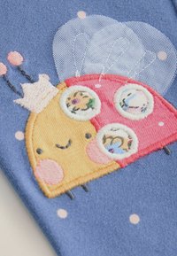 Broderad design av en leende skalbagge med gula och rosa sektioner, intrikata detaljer på kroppen, mot ett blått tyg med prickar.