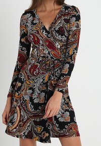 Mujer con un vestido envolvente de mangas largas, con una base negra y complejos patrones de paisley en rojo, naranja y blanco.