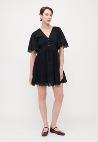 Schwarzes Baumwollkleid mit kurzen, ausladenden Ärmeln, V-Ausschnitt, Knopfleiste und Spitzenborte am Saum. Kombiniert mit burgunderfarbenen Ballerinas.