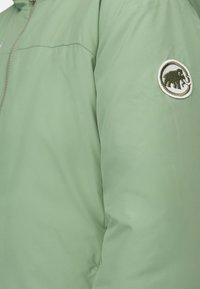 Giacca leggera verde con una texture liscia, chiusura con zip e un logo circolare di un elefante sulla manica sinistra.
