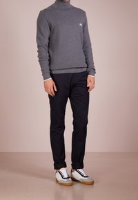 Pull à col roulé côtelé gris, pantalon noir et baskets blanches avec des accents gris et beige. Position debout sur un fond neutre.