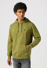 Sudadera con capucha verde oliva hecha de tela suave, con un bolsillo frontal y un pequeño logo en el pecho. Jeans grises visibles por debajo.
