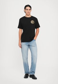 Svart bomulls t-skjorte med rund hals og et gyllent sirkulært logo på brystet, kombinert med lyseblå straight-leg jeans og svarte sko.