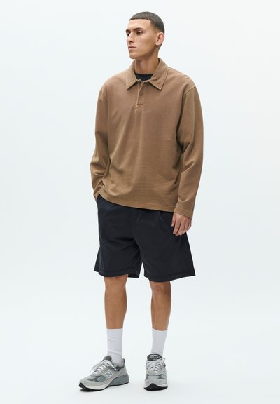 Barna hosszú ujjú galléros póló texturált felülettel, fekete rövidnadrággal és szürke sportcipővel párosítva.