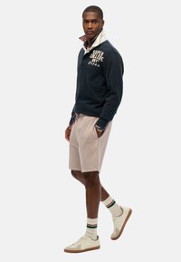 Pull marine avec un col écru et le texte "SUPER COLLEGE", associé à un short beige et des baskets blanches, avec des chaussettes rayées vertes.