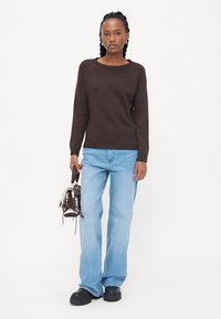 Brun strikket genser, romslig passform, rund hals. Lys blå rett jeans. Svarte platå sko. Holder svarte kunstskøyter.
