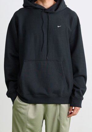 Person, die einen schwarzen Nike-Hoodie mit Fronttasche und Kordelzug trägt, kombiniert mit hellgrünen Hosen, vor einem einfarbigen hellen Hintergrund.